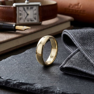5mm Slightly Domed Traditional Oval Wedding Band in 18K Yellow Gold, on walnut surface with pocket square