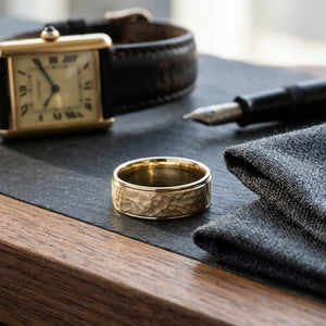 8mm Hammered Finish Carved Wedding Band in 18K Yellow Gold, on walnut surface with pocket square