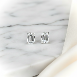 Emerald cut lab diamond stud earrings, four-prong basket, 18K white gold, on marble with silk